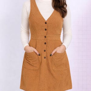 Woman wearing a brown corduroy Button Down Overall Dress with a white turtleneck and brown boots on a plain background.