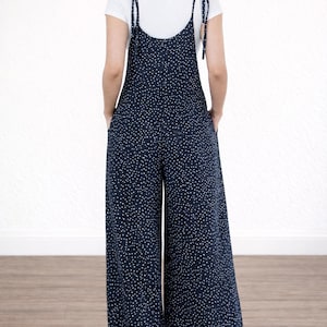 Back view of woman wearing navy Tie Shoulder Jumpsuit with white polka dots and white tee underneath on plain white background.