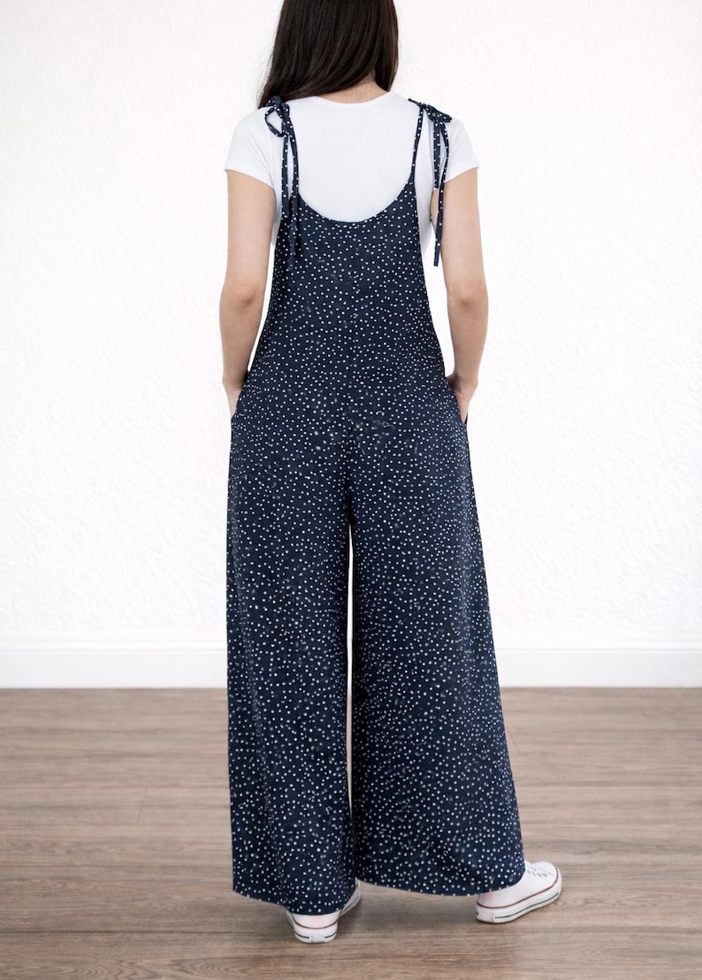 Back view of woman wearing navy Tie Shoulder Jumpsuit with white polka dots and white tee underneath on plain white background.