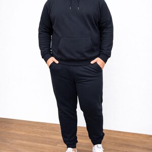 Person wearing a black Plus Size Mens Classic Hoodie and pants with white sneakers on a wooden floor.