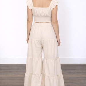 Back view of woman wearing beige top with ruffle shoulders and beige Boho Tiered Pants on wooden floor with plain white background.