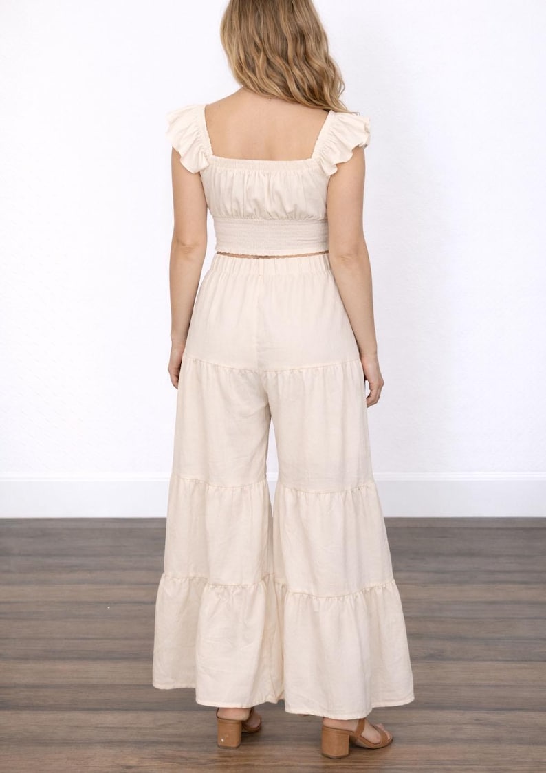 Back view of woman wearing beige top with ruffle shoulders and beige Boho Tiered Pants on wooden floor with plain white background.