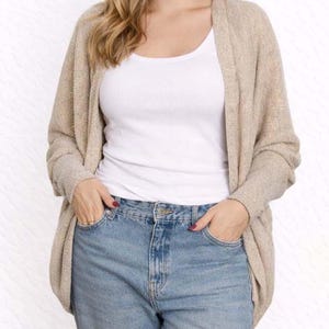 Woman wearing a beige Batwing Sleeve Cardigan, white tank top, and blue jeans on a wooden floor.