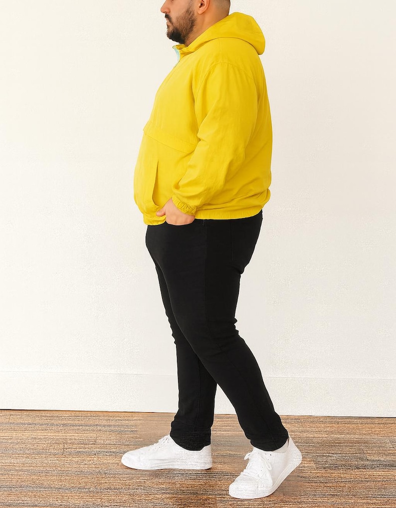 Side view of person wearing a yellow Plus Size Mens Windbreaker Jacket and black pants standing against a white wall.