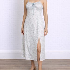 Woman wearing a white floral Milkmaid Midi Cami Dress with a tie neck and slit on a plain background.