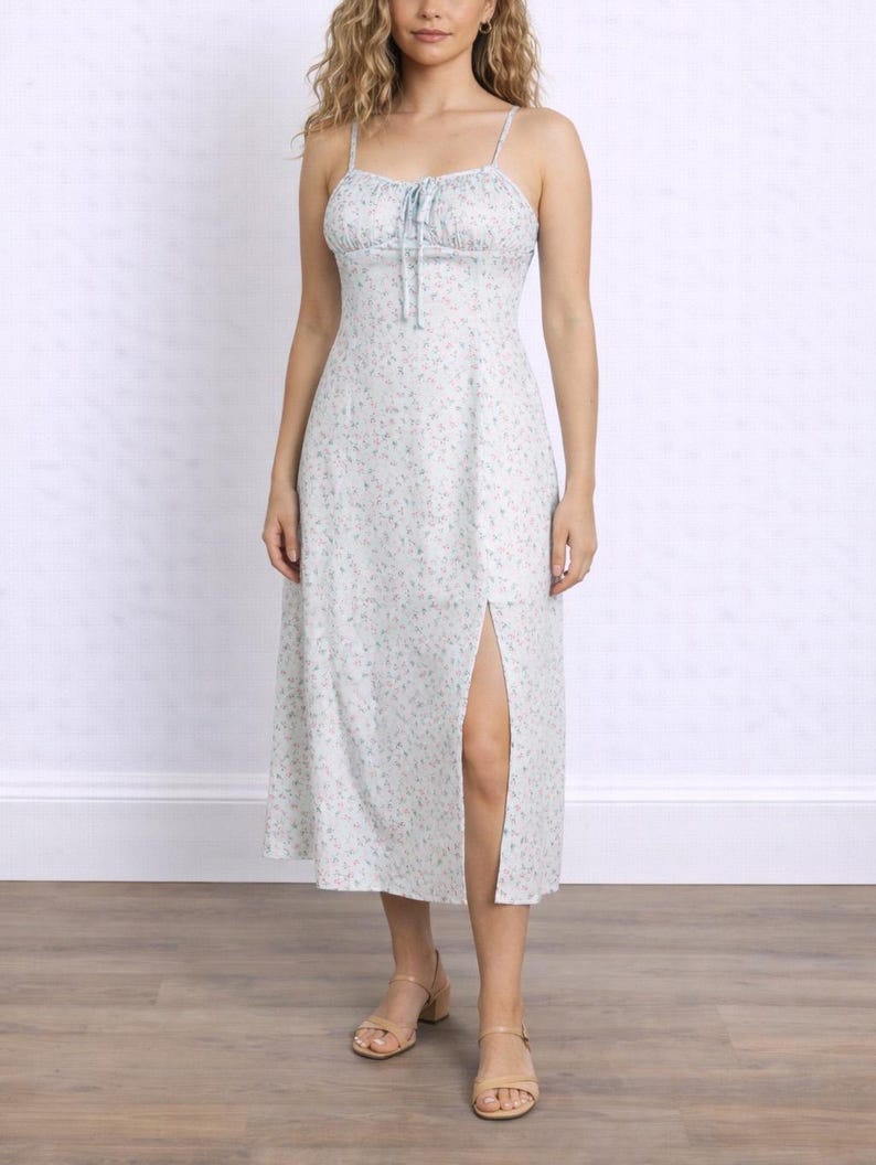 Woman wearing a white floral Milkmaid Midi Cami Dress with a tie neck and slit on a plain background.
