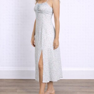 Side view of woman wearing a white floral Milkmaid Midi Cami Dress with a tie neck and slit on a plain background.