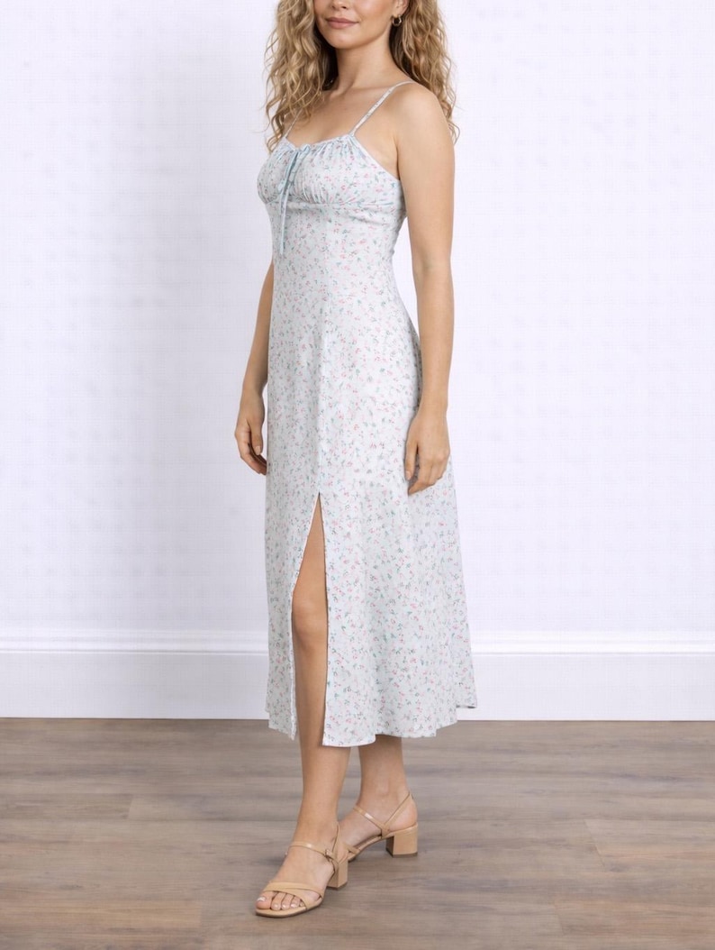 Side view of woman wearing a white floral Milkmaid Midi Cami Dress with a tie neck and slit on a plain background.