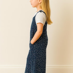 Side view of young girl wearing a navy blue polka dot Tie Shoulder Jumpsuit with white sneakers on a plain background.