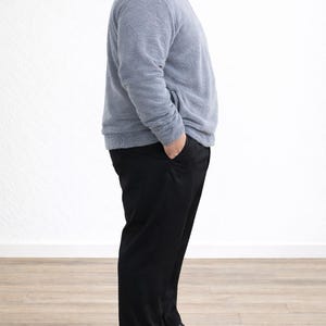 Side view of man wearing a gray Plus Size Mens Bomber Jacket and black pants standing against a white wall.