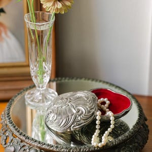 May include: A silver-toned metal trinket box with an ornate embossed lid and a vibrant red velvet interior lining. A strand of white pearls rests inside the open box. The box sits on a round, mirrored tray with a decorative dark metal base.
