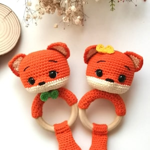 May include: Two crocheted fox rattle toys with wooden rings. The foxes are orange with white accents and have black eyes. One fox has a green bow tie and the other has a yellow bow.