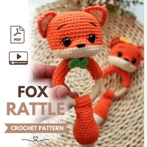 May include: A crochet fox rattle pattern featuring a bright orange fox head with black eyes, a small green bow tie, and a light wooden ring base. The text FOX RATTLE CROCHET PATTERN is displayed alongside icons for a PDF document and a video tutorial.