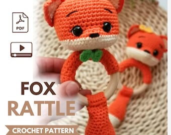 May include: A crochet fox rattle pattern featuring a bright orange fox head with black eyes, a small green bow tie, and a light wooden ring base. The text FOX RATTLE CROCHET PATTERN is displayed alongside icons for a PDF document and a video tutorial.