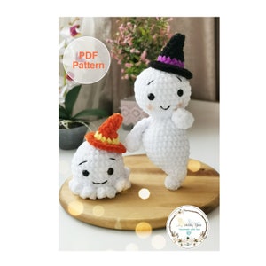 Könnte beinhalten: PDF-Muster zum Häkeln von zwei weißen Geister-Amigurumi mit orangefarbenen und violetten Hüten. Der größere Geist steht mit erhobenen Armen, während der kleinere Geist mit herabhängenden Armen sitzt. Beide Geister haben schwarze Augen und einen lächelnden Mund.
