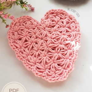 May include: A handmade, pink crocheted heart-shaped item with a textured floral pattern. The item is on a white surface, with the text "PDF Pattern" and "+ Video tutorials Step by step guide" visible. A gray crocheted item is in the background.