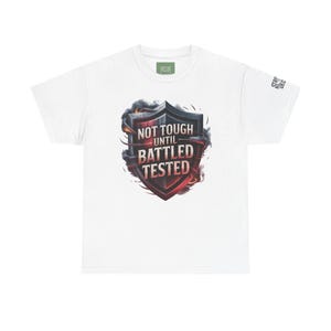 May include: White t-shirt with a shield graphic and the words "NOT TOUGH UNTIL BATTLED TESTED". The shield design uses dark and fiery colours with smoke effects. The shirt has a crew neck and short sleeves, a common style in the UK.
