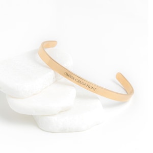 Puede incluir: Pulsera de brazalete de oro con la inscripción "Omnia Causa Fiunt".