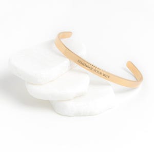May include: A gold-coloured cuff bracelet with the engraved text "REMEMBER YOUR WHY". The bracelet is resting on a stack of white, smooth stones. The bracelet has a simple, open design.