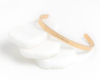May include: A gold-coloured cuff bracelet with the engraved text "I AM ENOUGH" is displayed on a white background. The bracelet is resting on top of three white, smooth, irregularly shaped stones. The bracelet has a simple, minimalist design.