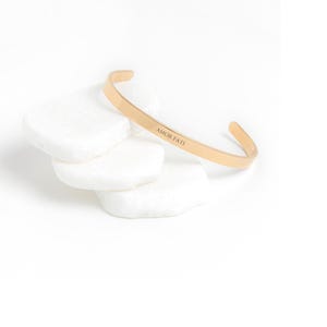 May include: A gold-toned bangle bracelet with the inscription "AMOR FATI" on a white background.