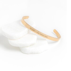 May include: Gold-toned bangle bracelet with the inscription "Progress Over Perfection" engraved on the band.