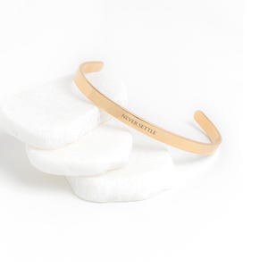 May include: Gold-toned bangle bracelet with the inscription "NEVER SETTLE" on a white background.