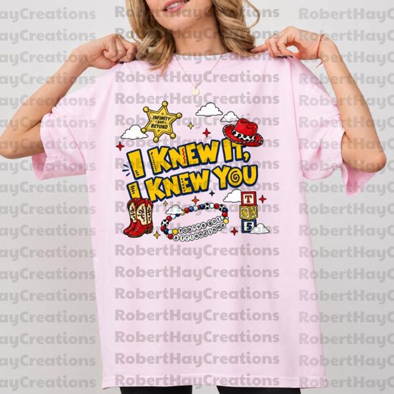 Puede incluir: Una camiseta rosa claro con un estampado gr&aacute;fico que incluye el texto I KNEW IT, I KNEW YOU. El dise&ntilde;o presenta una estrella de sheriff amarilla, un sombrero de vaquero rojo, una bota de vaquero roja, una pulsera de cuentas y tres cubos.
