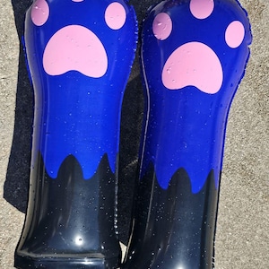 May include: Two inflatable blue paw-shaped pool floats with pink paw prints. The bottom portion of the floats is black. The floats are lying on a concrete surface.