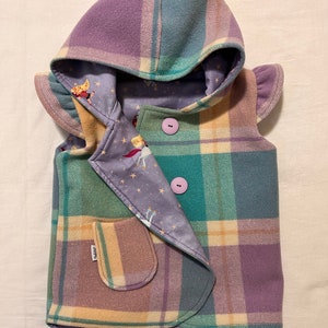 May include: A hooded vest with a plaid pattern in shades of purple, green, and cream. The vest has a pocket on the front and two buttons. The lining of the vest is a light purple fabric with a star and unicorn print.
