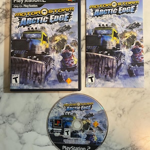 Puede incluir: Un videojuego de PlayStation 2, "MotorStorm Arctic Edge", con su caja, carátula y disco. La carátula presenta un vehículo amarillo y azul compitiendo en un entorno nevado. El disco del juego es plateado con el título y el logotipo.