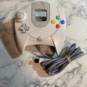 Puede incluir: Un mando de Dreamcast blanco y gris con un cable gris y un conector negro. El mando tiene una cruceta, una palanca analógica y botones de acción de colores. También se ve un botón de inicio.