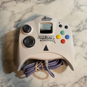 Puede incluir: Mando Dream Shock blanco con cable morado. El mando tiene una cruceta, un stick analógico y botones de colores. El texto "DREAM SHOCK" es visible en el mando.