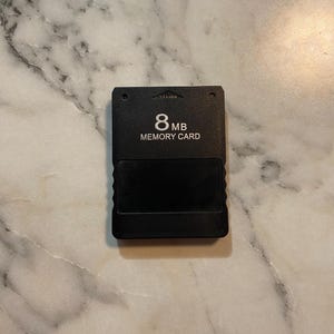 May include: A black 8 MB memory card with the text "8 MB MEMORY CARD" printed on the front. The card has a rectangular shape with a slot at the bottom and a small arrow at the top.