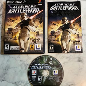 Puede incluir: Un conjunto de videojuegos para PlayStation 2 "Star Wars: Battlefront". La caja y el disco del juego muestran un Stormtrooper con un blaster, con el título en un lugar destacado. El juego está clasificado como "Teen".