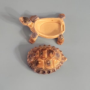 Peut inclure: Un vide-poche en céramique en forme de tortue avec une carapace amovible. Le plat est beige avec des détails bruns sur la tête, les pattes et la carapace. La carapace est une pièce séparée, avec un motif texturé détaillé.