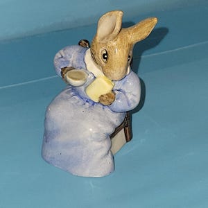 May include: A ceramic figurine of a rabbit in a blue coat, holding a yellow object. The rabbit has brown fur and is on a light blue surface. The figurine is a collectible item, possibly from a children's story.