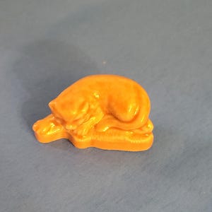 May include: A small, orange cat figurine in a sleeping position. The cat is curled up with its head down and paws extended. The figurine is made of a glossy material and sits on a textured base. The cat is a solid orange colour.