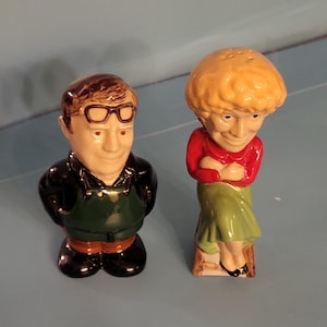 May include: A pair of ceramic salt and pepper shakers. One is a man wearing glasses, a black jacket, and a green apron. The other is a woman in a red top and green skirt. Both have exaggerated features.