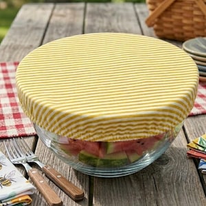 May include: A clear glass bowl filled with watermelon slices, covered with a yellow and white striped fabric bowl cover. The bowl sits on a wooden picnic table, with a red and white checkered cloth and silverware.