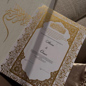 May include: A white marriage certificate featuring gold foil calligraphy and intricate borders. The text displays the names Amna and Omar, alongside Islamic verses. The document is shown with a matching white folder featuring gold Arabic script.