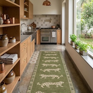 May include: A green runner rug with a repeating leopard pattern in a kitchen setting. The rug is rectangular with a cream-colored border and fringe. The kitchen features wooden cabinets, copper light fixtures, and a large window.