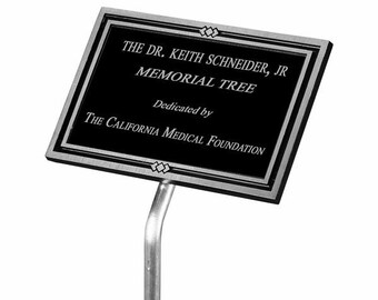 May include: A black metal plaque with silver trim and a white inscription that reads "THE DR. KEITH SCHNEIDER, JR MEMORIAL TREE Dedicated by THE CALIFORNIA MEDICAL FOUNDATION". The plaque is mounted on a silver metal stand.