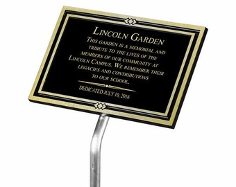 May include: A rectangular black sign with gold trim and a silver post. The sign reads "LINCOLN GARDEN" and includes text about the garden's purpose and dedication date of July 10, 2016. The sign is for a memorial at Lincoln Campus.