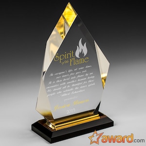 Puede incluir: Un premio de cristal transparente con un borde dorado y una base negra. El premio presenta un diseño de llama y el texto "Spirit of the Flame". El premio se entrega a Jessica Bowers en 2015.