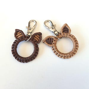 Op de afbeelding: Twee gebreide sleutelhangers in bruin en beige met een ring en een zilveren metalen sluiting. Elke sleutelhanger heeft twee kleine oorvormige lussen.