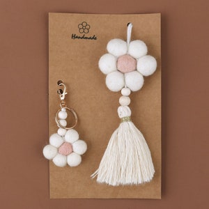 May include: A handmade set of two daisy-shaped accessories. One is a keychain with a gold clasp, and the other is a tassel with wooden beads. Both have white felt petals and a pink center, displayed on a brown card.