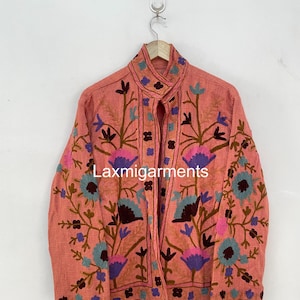 Pode incluir: Casaco coral com bordados florais intrincados em tons de azul, roxo e castanho. O casaco apresenta uma gola alta e está pendurado num cabide de madeira. O texto "Laxmigarments" é visível no casaco.