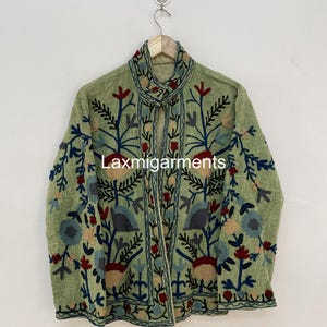 Pode incluir: Casaco verde claro com bordados florais intrincados em tons de azul, vermelho e bege. O casaco tem uma gola alta e é exibido em um cabide de madeira. O texto "Laxmigarments" é visível no casaco.