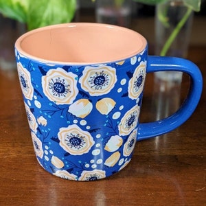May include: A blue ceramic mug with a floral pattern. The mug has a peach-coloured interior and a white floral design on a blue background.